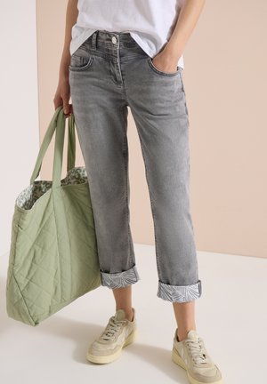 Personne portant un jean gris retroussé, des baskets blanches, une chemise blanche, tenant un grand sac cabas vert matelassé avec une doublure à motifs.