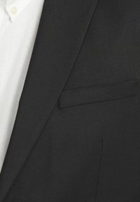 Blazer noir avec un revers structuré, détail de poche unique et texture fine. En dessous, une chemise blanche avec un col boutonné est visible.