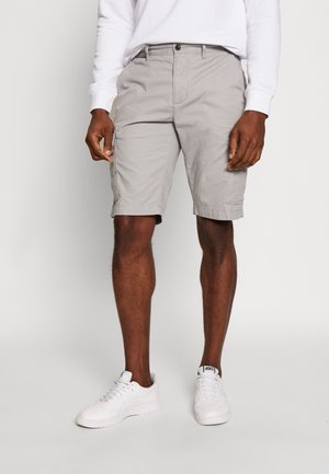 Homme portant un short cargo gris clair et des baskets blanches, debout devant un fond uni clair.