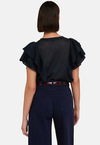 Granatowa bluzka z falbaniastymi rękawami i teksturowanym materiałem, zestawiona z dopasowanymi ciemnymi spodniami i błyszczącym brązowym paskiem. Widok z tyłu.