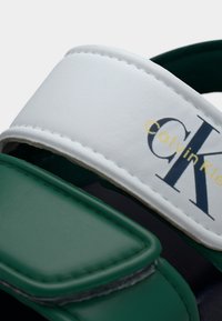 Κοντινή λήψη πράσινων και λευκών λουριών σανδάλων με το λογότυπο Calvin Klein τυπωμένο στο λευκό λουρί.