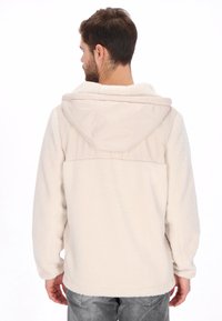 Chaqueta de forro polar color crema con capucha y panel en los hombros de tela lisa beige. Presenta un ajuste holgado y mangas largas.