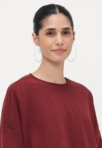 Sudadera burdeos con escote redondo, hombros caídos y textura suave. Un modelo lleva pendientes de aro plateados y un maquillaje minimalista.
