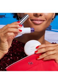 Tre skönhetsprodukter: ett silverläppstift, en vit penna med en lila topp och en vit kompakt med etiketten "KIKO", som hålls ovanför en röd paljetväska.