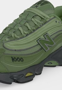 Πράσινο αθλητικό παπούτσι New Balance 1000 με πλεκτό ανώτερο μέρος, χονδρές κορδόνια και μαύρη σόλα με επένδυση ορατή μέσα από το πλαϊνό παράθυρο.