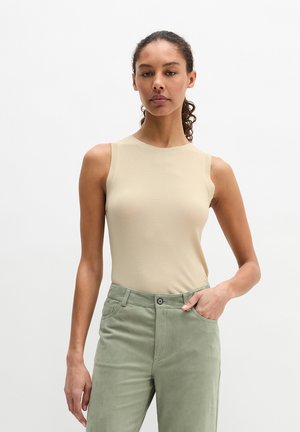 Femme portant un haut beige sans manches et un pantalon vert clair, debout avec une main dans la poche, devant un fond blanc uni.