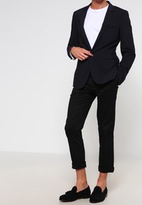 Man i svart randig blazer, vit t-shirt, svarta uppvikt byxor och svarta slip-on loafers, stående mot en enfärgad bakgrund.