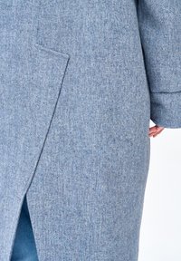 Zbliżenie na jasnoniebieski, teksturowany płaszcz wełniany z rozcięciem z tyłu, widoczna ręka modelki po boku.