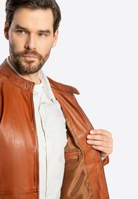 Braune Lederjacke mit glatter Textur, ausgestattet mit einem Reißverschluss, Druckknöpfen und einer Innentasche. Leichtes Design.