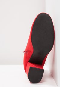 Czerwone zamszowe szpilki z kwadratowym noskiem, teksturowanym obcasem i czarną gumową podeszwą. Szwy wzdłuż boku dodają wizualnego zainteresowania.