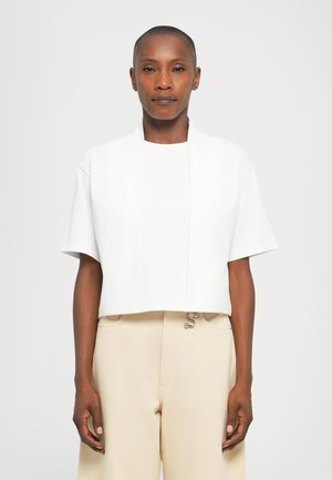 Person with short hair wearing a white cropped open jacket over a white t-shirt and beige high-waist pants, standing against a plain background.