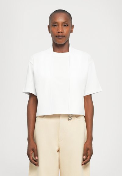 Persona con capelli corti che indossa una giacca bianca corta aperta su una t-shirt bianca e pantaloni beige a vita alta, in piedi contro uno sfondo uniforme.