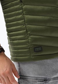 Ciemnozielona pikowana kamizelka z pionowymi przeszyciami, teksturowaną powierzchnią oraz czarną metką z zielonym napisem na dolnej stronie.