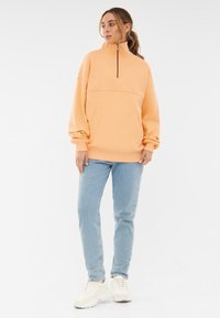 Oranje sweatshirt met een hoge kraag, een halve rits aan de voorkant en zijzakken. Gekleed met lichtblauwe jeans en witte sneakers.