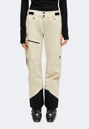 Personne portant un pantalon de ski beige avec le bas des jambes noir, des poches zippées noires et des chaussures de ski noires, debout devant un fond uni.