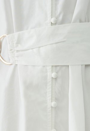 Biele šaty s textúrovanou látkou, širokým opaskom v páse, veľkým okrúhlym zlatým kovovým doplnkom a jemnými gombíkovými detailmi na prednej strane.