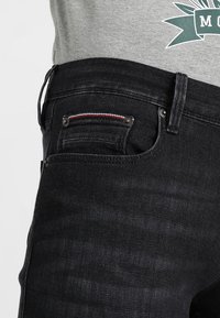 Vaqueros de mezclilla negra con un ajuste entallado, que cuentan con un pequeño bolsillo frontal con costuras rojas y azules, botones de metal y una textura suave.