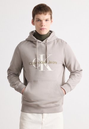 Light tan hooded sweatshirt with white and gold Calvin Klein Jeans logo. Features a kangaroo pocket and ribbed cuffs at the sleeves.