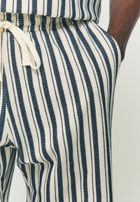 Pantalons rayés en tissu avec des lignes alternées bleu marine et blanc cassé. Présentent une taille élastique avec un cordon de serrage et des poches latérales.