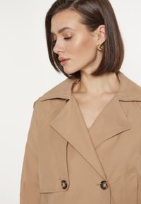 Trench coat bej cu două rânduri de nasturi, având buzunare mari în față și rever cu decupaje, cu nasturi lucioși și o textură netedă.