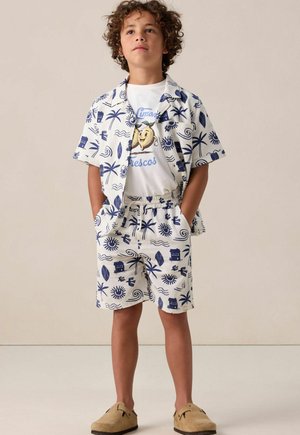 Korte mouwen witte shirt met marineblauw tropisch patroon, bijpassende shorts en een witte T-shirt met een gele fruitafbeelding. Bruine instap schoenen.