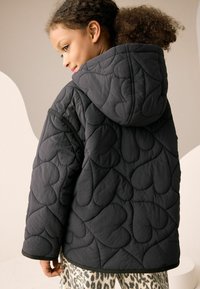 Veste noire matelassée avec une capuche, présentant un motif de cœur, un tissu texturé et une coupe décontractée. Soulignée par un bord noir le long de l'ourlet.