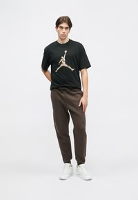 Schwarzes T-Shirt mit einem bunten Grafikan design eines Basketballspielers, kombiniert mit braunen Jogginghosen und weißen Turnschuhen.