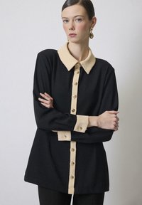Zwarte blouse met lange mouwen en een beige kraag en knoopsluiting, voorzien van knopen in goudkleur en een ruime pasvorm. Glad stoftextuur.