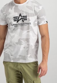 Biała koszulka z kamuflażem, wykonana z bawełny, z czarnym logo i napisem "ALPHA INDUSTRIES". Krótkie rękawy i okrągły dekolt.