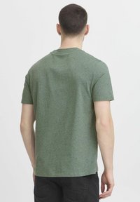 Camiseta de manga corta verde fabricada con tela texturada con un patrón moteado. Cuello redondo y un ajuste holgado. Vista trasera.