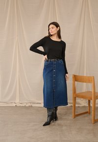 Moteris juodu ilgarankoviu palaidine, susagstoma mėlynos džinsinės midi sijonu, juodais aukštakulniais batais, stovinti šalia medinės kėdės ant betoninio grindinio.