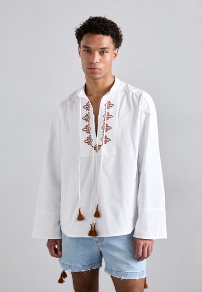 Chemise blanche à manches longues avec une broderie brune décorative le long du col et des franges à l'ourlet, assortie à un short en denim.