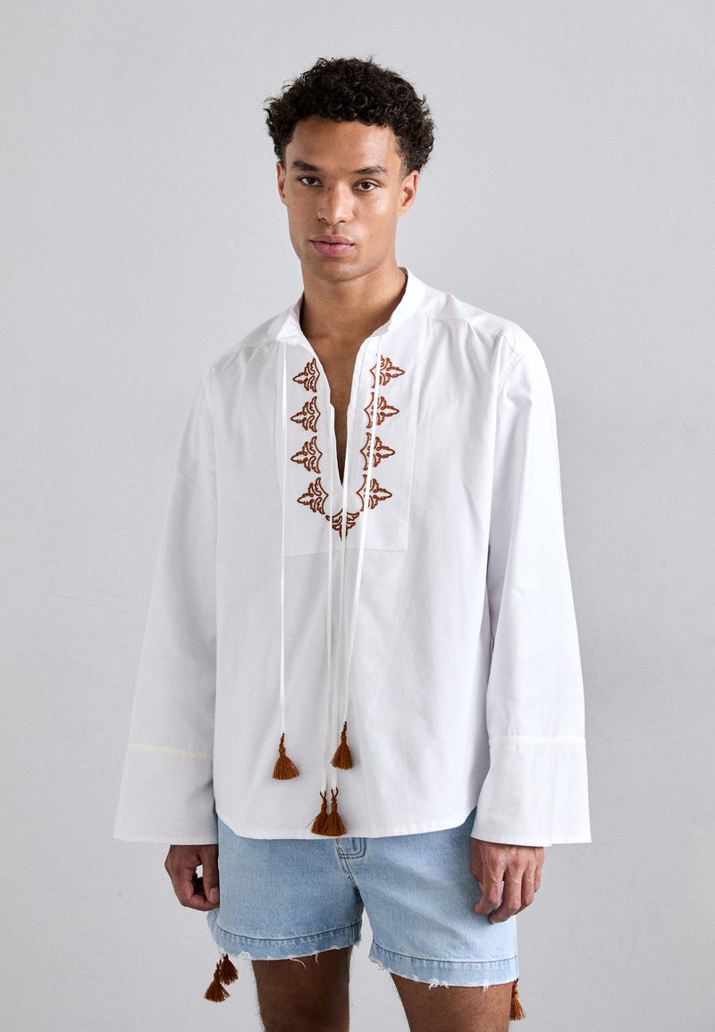 Chemise blanche à manches longues avec une broderie brune décorative le long du col et des franges à l'ourlet, assortie à un short en denim.