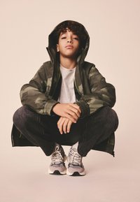 Žalia kamufliažinė striukė su gaubtu, pilka marškinėliai, tamsūs džinsai ir pilki sportbačiai su šviesiomis detalėmis. Modelis prisikūprinęs, rankos remiasi į kelius.