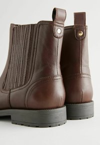 Botas de tobillo de cuero marrón con paneles laterales elásticos, puntera redondeada y suela de goma plana. Presenta acentos de hardware en tono dorado.