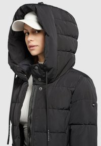 Sort pufferjakke med stor, polstret hætte, frontlynlås og snore. Har quiltet tekstur og et logo på ærmet.