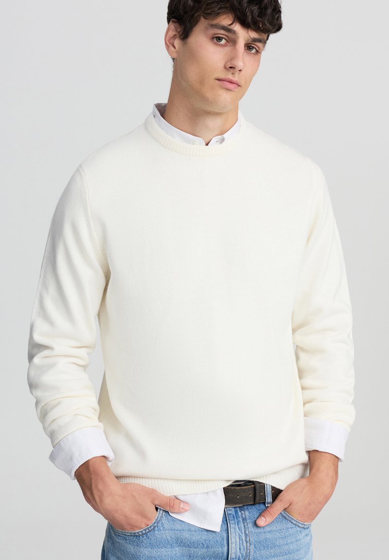 Maglione a maglia color crema con colletto alto e polsini a coste, abbinato a una camicia bianca con il colletto visibile sotto. Tessuto texturizzato.