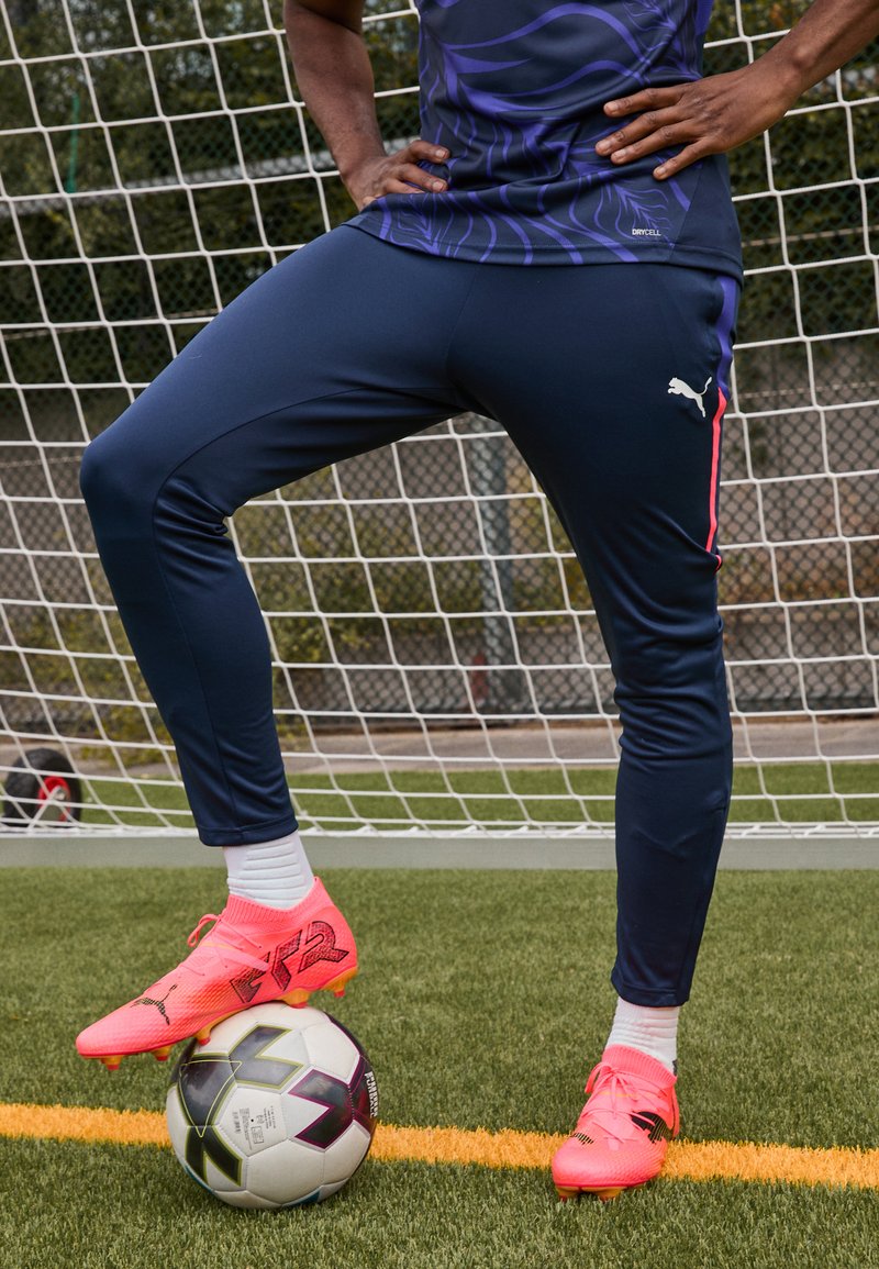Puma sporta apģērbs, kas ietver pieguļošas tumši zila bikses ar rozā sānu akcentiem un vieglus rozā futbola zābakus, stāvot uz melnbaltā futbola bumbas.
