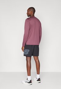 Langærmet maroon skjorte med rund hals, parret med sorte shorts med et blåt "NB" logo. Modellen bærer hvide sneakers med sorte detaljer.