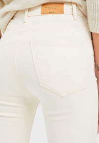 Jeans de denim blanco con un diseño ajustado, presentan una etiqueta de cuero en la parte posterior y un bolsillo trasero estándar. Tejido texturizado con un ligero brillo.