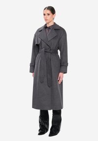 Grå uld trenchcoat med bred krave, taljebælte, dobbeltradet front og opkørte manchetter, kombineret med sorte, tætsiddende knæhøje støvler.