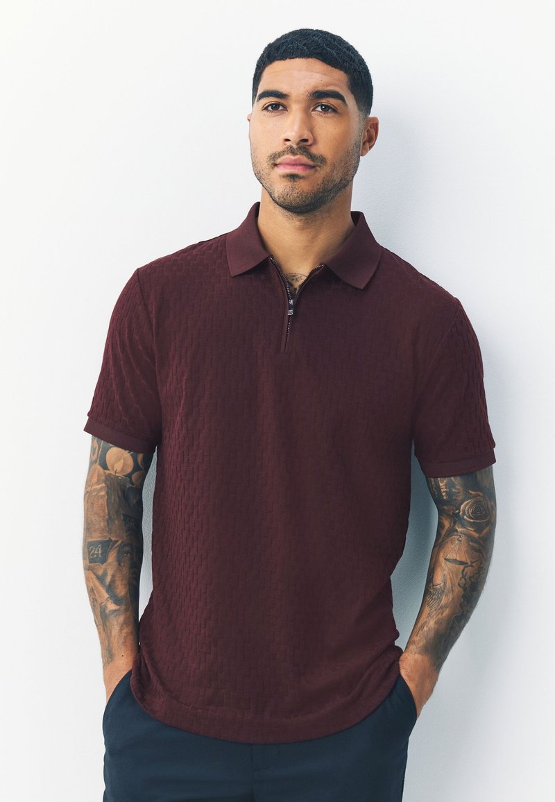 Polo bordeaux con collo a zip e motivo a trama; maniche corte; abbinata a pantaloni scuri. Presenta una vestibilità slim e una lucentezza discreta.