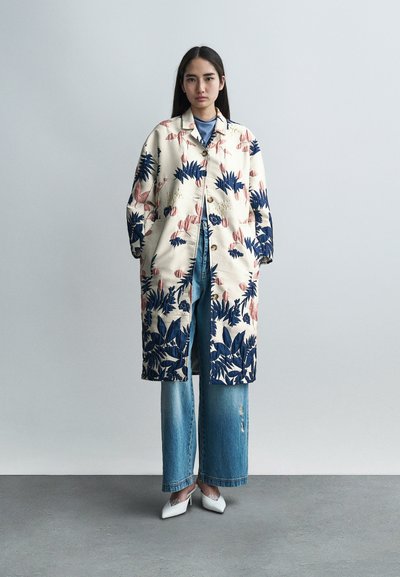 Manteau long crème avec des motifs floraux et de feuilles en marine et rose. Associé à un haut bleu clair, un jean large déchiré et des talons blancs.