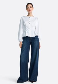 Biela blúza s dlhým rukávom, rovný výstrih a zapínanie na gombíky, v kombinácii so širokými tmavými džínsami. Jednoduchý, štruktúrovaný dizajn.