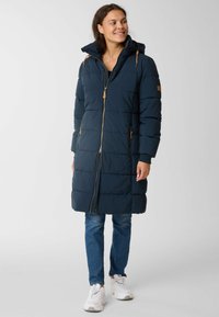 Lange marineblauwe gewatteerde jas met een rits aan de voorkant, tan ritsaccenten, een capuchon en zijzakken, gedragen over blauwe jeans en witte sneakers.