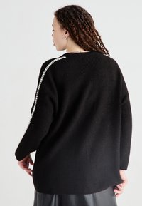 Czarny, oversize sweter z ryżowym wzorem, z białymi paskami przy ramionach. Miękki, luźny krój, dłuższy tylny dół.