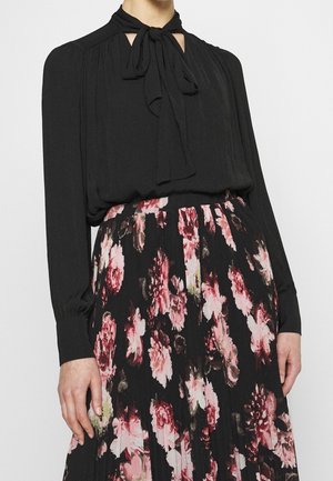 Femme portant un chemisier noir à manches longues avec cravate et une jupe plissée noire à motif floral rose et rouge.