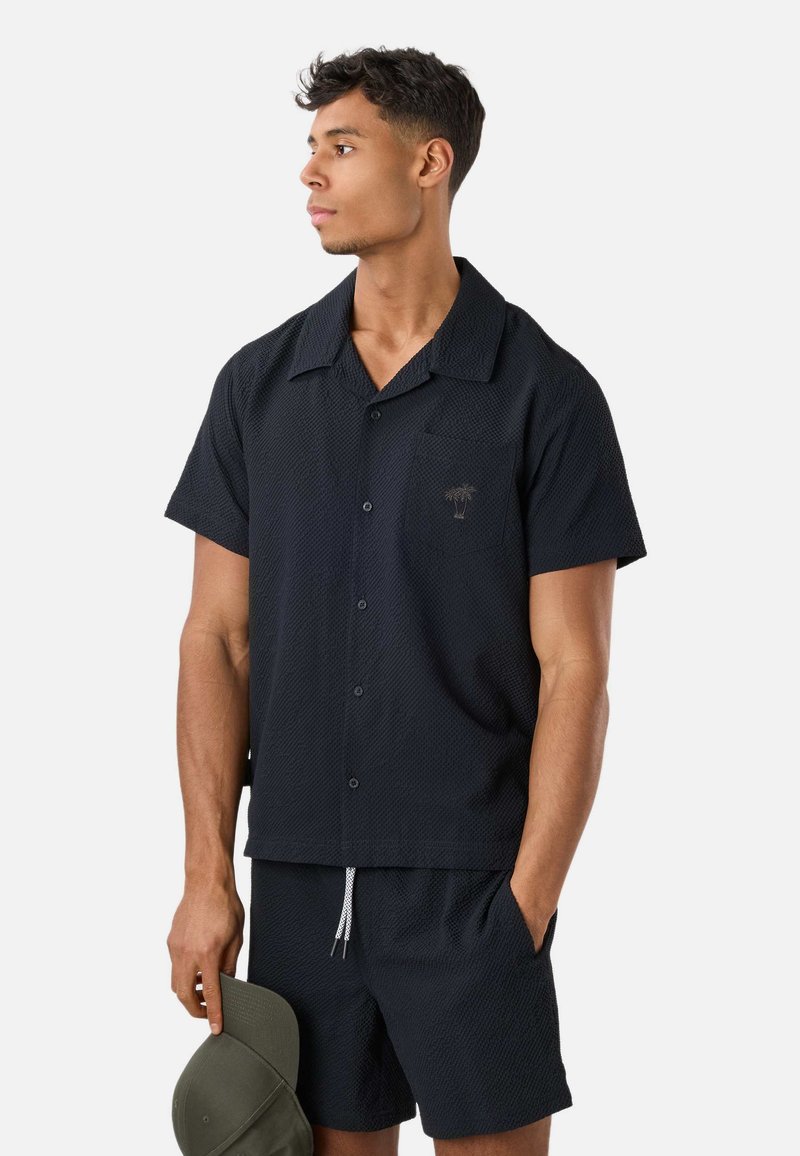 Jeune homme regardant à gauche portant une chemise noire à manches courtes boutonnée et un short assorti, tenant une casquette vert olive dans une main.