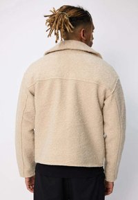 Veste beige en mélange de laine avec un grand col, un tissu texturé et des coutures aux épaules. Vue de dos montrant une coupe décontractée et une construction simple.