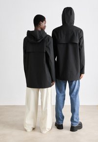 Două persoane poartă jachete cu glugă negre, cu un design cu clapetă pe spate. Una are pantaloni largi cremoși, iar cealaltă blugi și cizme negre.